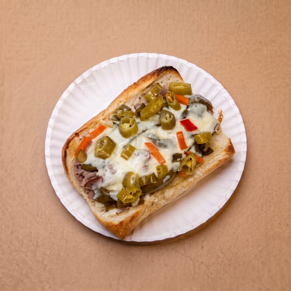 Melted cheese and pickled peppers on a sub sandwich served on a white paper plate.