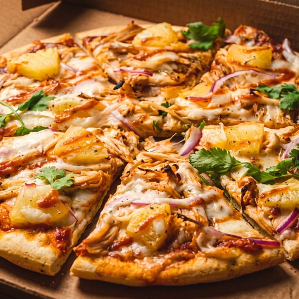 Sliced BBQ chicken and pineapple pizza with red onion and cilantro in a cardboard box.