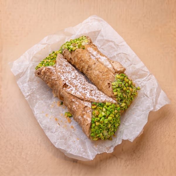 Two cannoli with powdered sugar and pistachio pieces on white parchment paper.