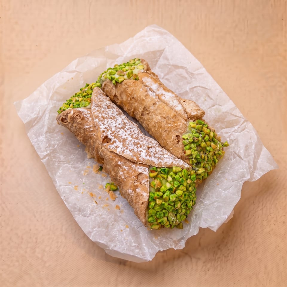 Two cannoli with powdered sugar and pistachio pieces on white parchment paper.